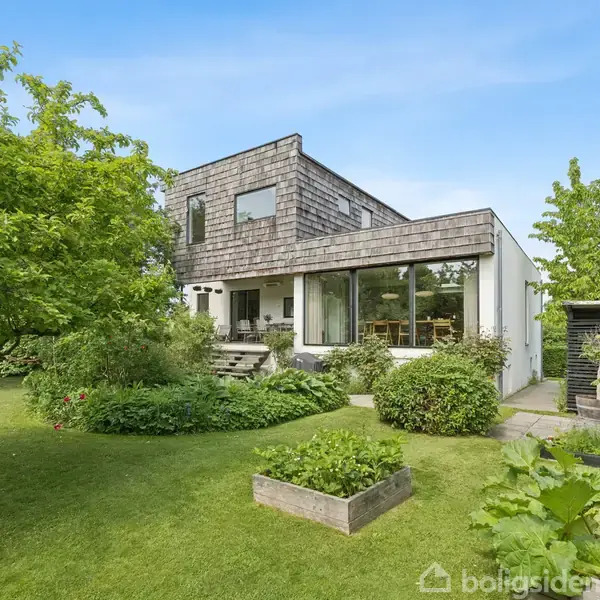 Et moderne hus med glaspartier og træfacade står i en frodig have med grøn græsplæne, buske og træer. Terrassen er møbleret, og omgivelserne er grønne og velplejede.