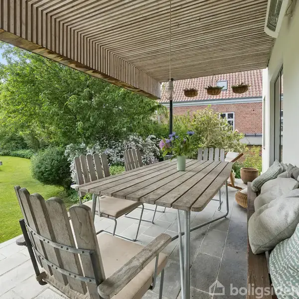 Lang træbord omgivet af stole står på en terrasse med tæppe og puder, mens træer og græsplæne omkranser huset i baggrunden.