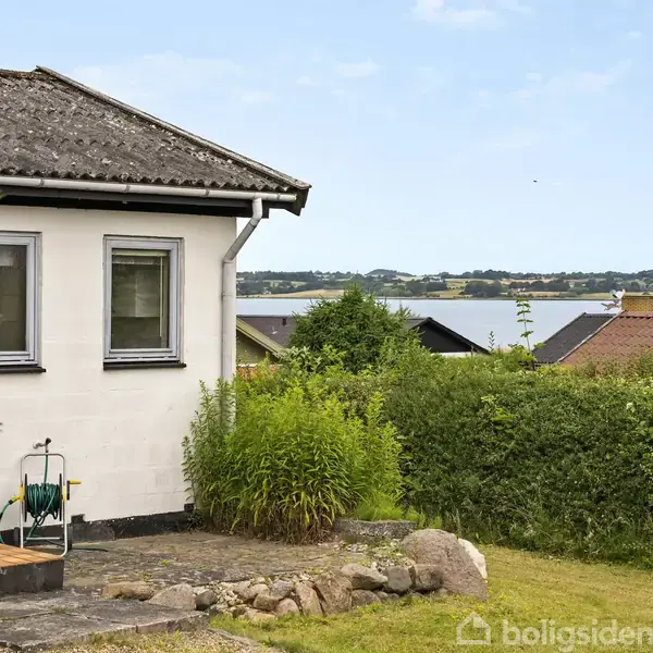 Et hvidt hus med tækket tag står i en have med buske og sten. I baggrunden ses en udsigt over vandet og et landskab med huse og træer.