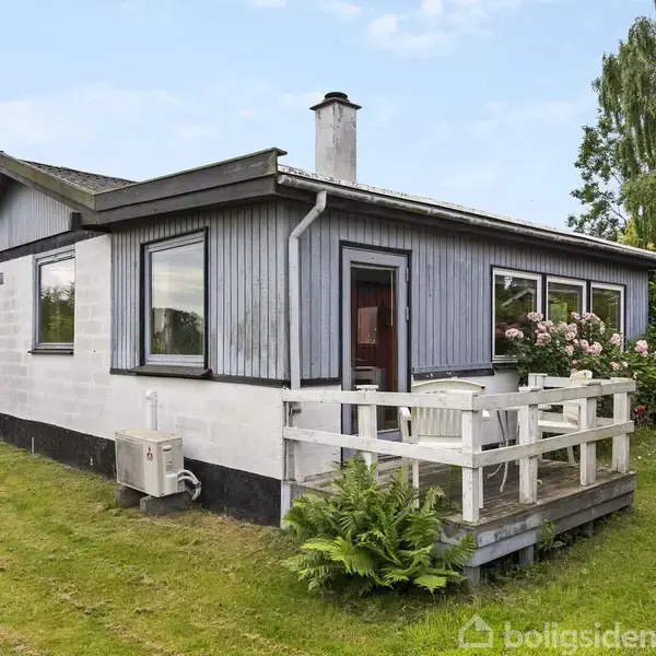 Et lille sommerhus står på en græsplæne. Huset har en terrasse med hvide møbler og blomster. Omgivet af træer og buske i et grønt, hyggeligt haveområde.