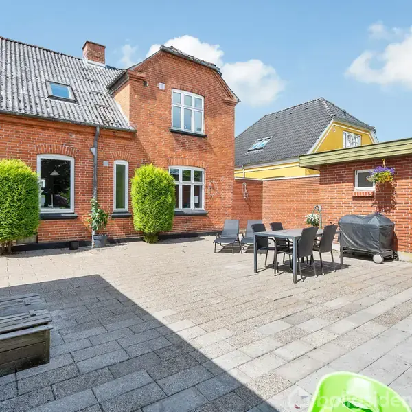 Et rødt murstenshus med skråt tag omkranser en gårdhave. En terrasse af fliser har et bord med stole og en grill i baggrunden. Cykler og planter står langs væggene.