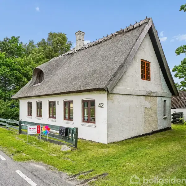 Et stråtækt hus med hvide vægge står ved en landevej. Husnummer 42 er tydeligt vist. Omgivet af grønne træer og græsplæne i et landligt miljø.