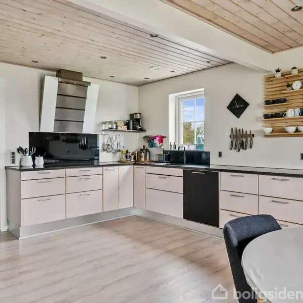 Lyst køkken med hvide skabe og moderne apparater, ser ud mod et vindue. Et rundt bord står i forgrunden, mens hylder med kopper og glas hænger på væggen.