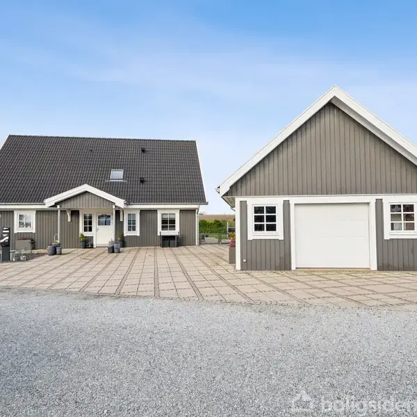 En gråmalet træhus med skråt tag står stille ved siden af en separat garage. De er omgivet af flisebelagt indkørsel og havehæk under en klar, blå himmel.