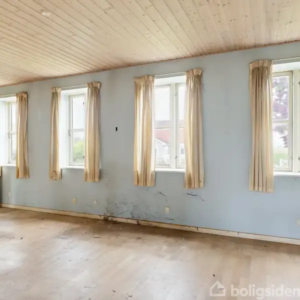 Rum med trægulv og loft, hvide vægge, fire vinduer med beige gardiner. Der er synlige fugtskader ved væggen. Åben dør i hjørnet med adgang til et tilstødende rum.