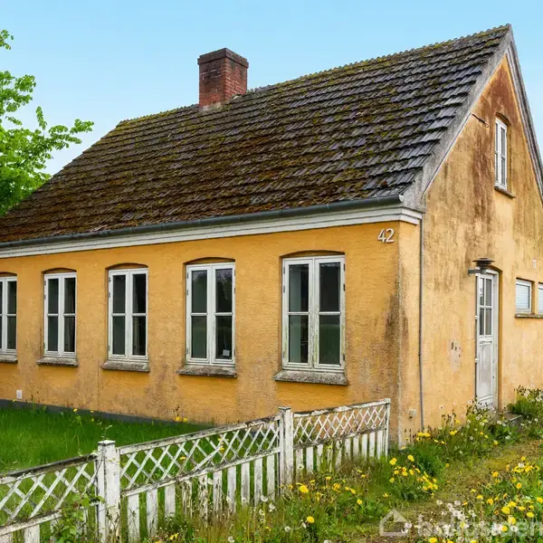Et gulmalet hus med fire vinduer og skifertag står i en have med en lav, hvid stakit. Nummer 42 er tydeligt synligt på facaden. Baggrunden har grønne træer.