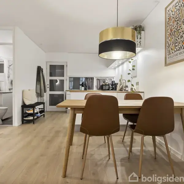 Spisebord med fire stole står under en sort og guld lampe i en moderne spisestue. Til venstre er der et åbent badeværelse, og et farverigt billede hænger på væggen.