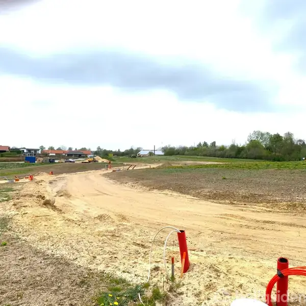 Sandet vej fører gennem en mark med rød-orange markeringsstokke, afsluttende ved en landsby med huse i baggrunden. Himlen er overskyet, og området er sparsomt grønt.