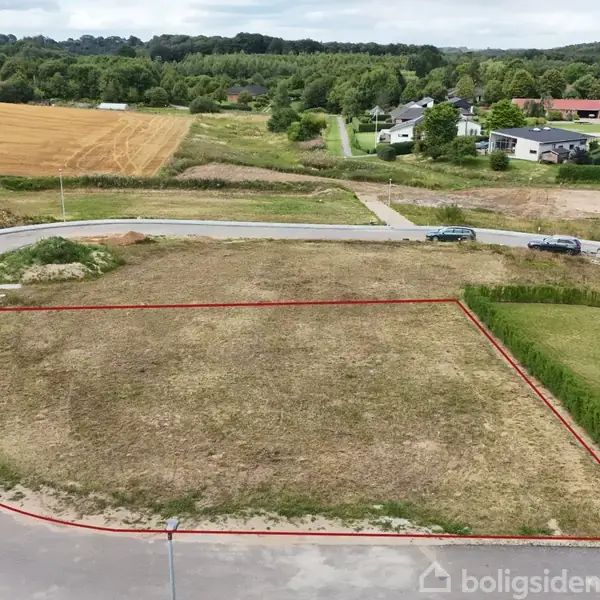 Grundstykke markeret med en rød linje ligger ved en vej i et boligområde. Småhuse og træer ses i baggrunden, omgivet af marker og grønne områder.