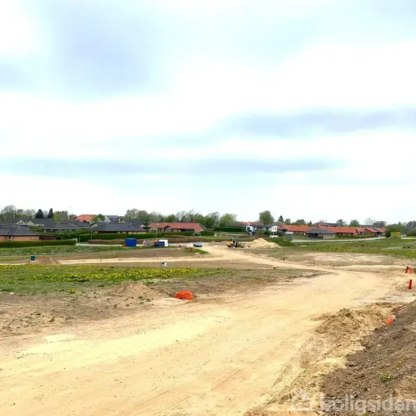 En byggeplads viser en jordvej omkranset af grøn natur og spredte huse i baggrunden under en overskyet himmel. Ingen synlig aktivitet bemærkes på byggepladsen.