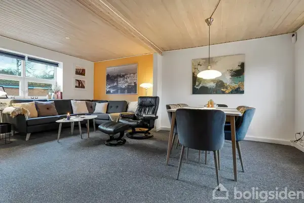 Stuen består af en grå sofa med farverige puder, en lænestol med skammel, et spisebord med fire stole. Store vinduer og lys vægudsmykning skaber en hyggelig atmosfære.