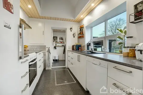 Et moderne køkken med hvide skabe og sort bordplade. Køkkenet har en ovn, opvaskemaskine og vask. Store vinduer giver udsigt til haven udenfor. Lyse detaljer på loftet skaber en rummelig atmosfære.