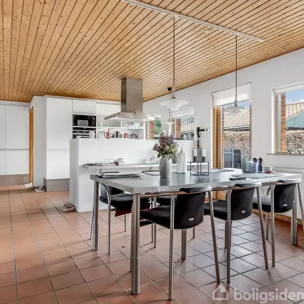 Et moderne køkken-alrum med spisebord i midten omgivet af sorte stole. Lysindfald fra vinduerne. Køkkenet har hvide skabe og flisegulv. Loftet er beklædt med træ.