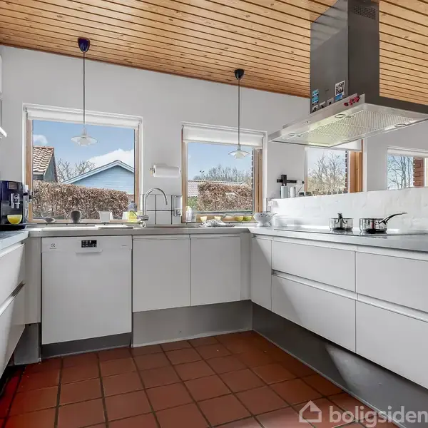 Køkken med hvide skabe og stålbordplader; pander står på komfuret under emhætten. Store vinduer med udsigt til have. Rødt flisegulv og træloft skaber en varm atmosfære.