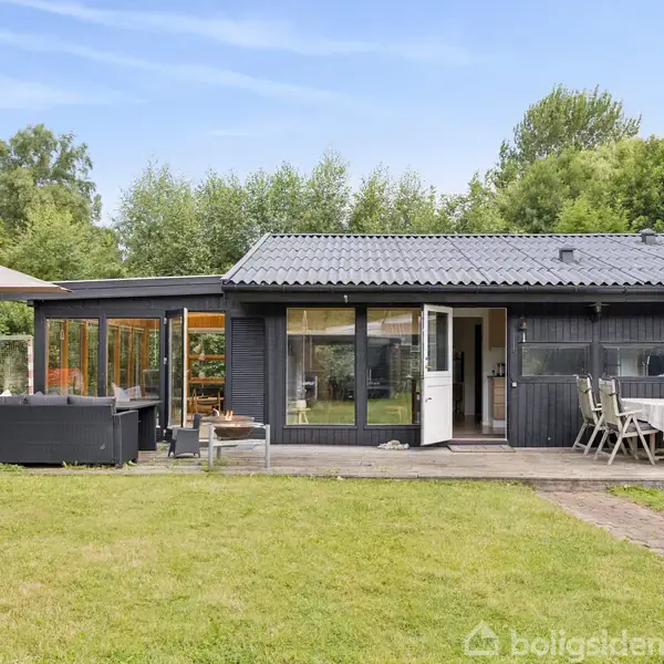 Et sortmalet sommerhus med åbne døre ses på en græsplæne med havemøbler og en parasol. Omgivet af grønne træer skaber det en rolig, naturlig atmosfære.