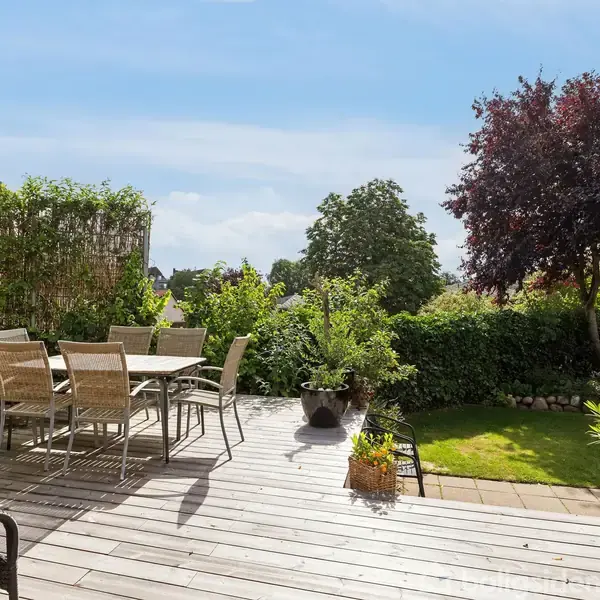 Terrasse med metal- og glasbord samt stole står under en stor parasol. Omgivet af grønt buskads og træer i en have på en solrig dag.