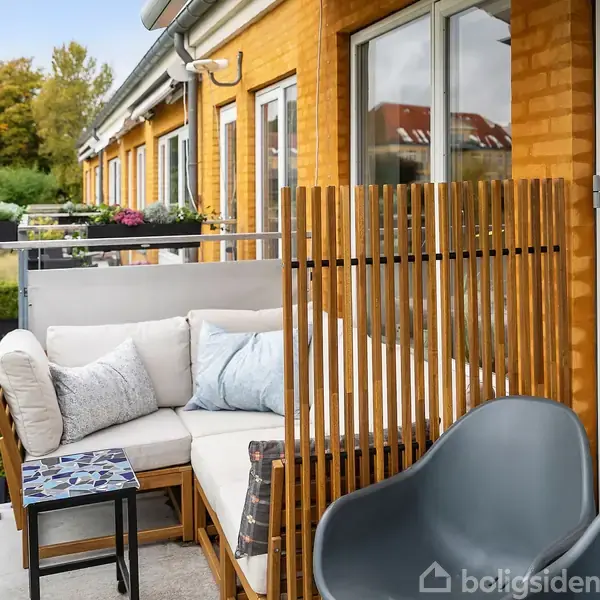 En hyggelig altan med en hvid sofa, puder, lille bord og to grå stole, omgivet af et glasrækværk. Siden af huset er gul mursten. Træer ses i baggrunden.