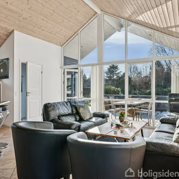 Sort lædermøblement står i en stue med højt til loftet, store vinduer giver udsigt til en solrig terrasse og have. Trægulv og loft skaber en lys, rummelig atmosfære.