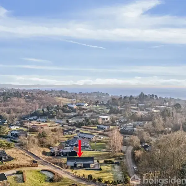 Et hus markeret med en rød pil ligger i en landsby omgivet af træer og andre huse. Udsigten viser et kuperet landskab under en klar himmel.