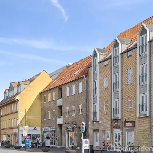 Flere forskelligfarvede bygninger står side om side på en solrig gade. Nogen har butikskilte som "Czyz Kiosk" og andre som boligindgange. Blå himmel i baggrunden.