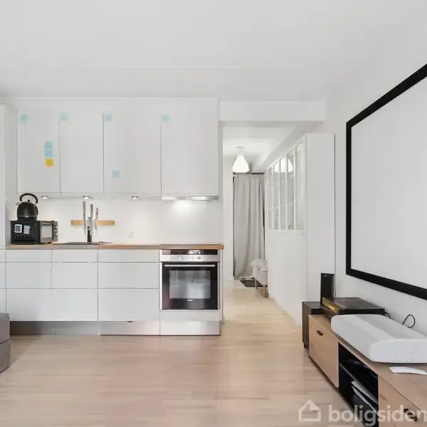 Et lyst køkkenområde har en hvid sofa ved siden af. Der er et sort-hvidt indrammet panel på væggen. Til højre er der et lille skab ved siden af en projektor.