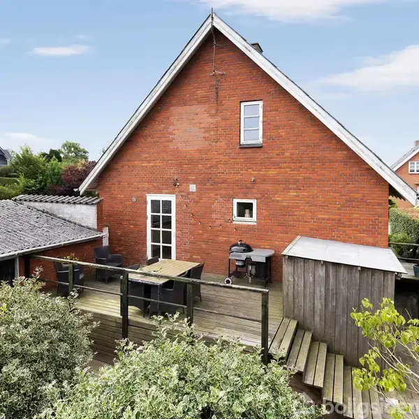Rødt murstenshus med trekantet tag, placeret tæt ved en træterrasse med grill og bord. I baggrunden ses en vej og flere huse. Grønt buskads omgiver terrassen.