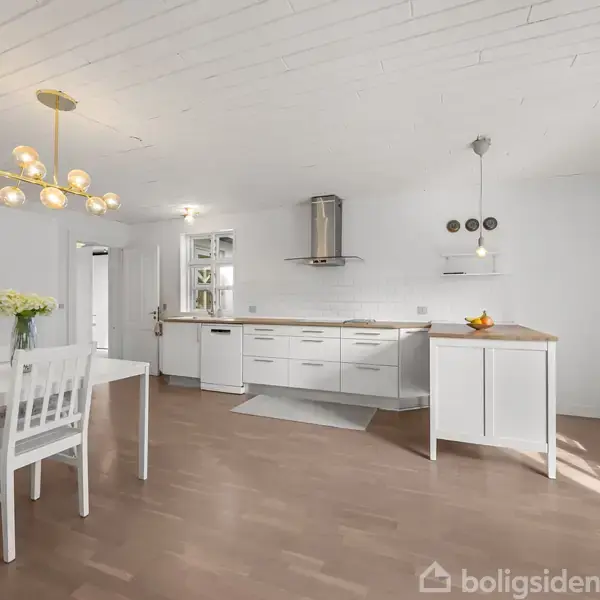 Køkken med hvide skabe og lys træbordplade. Et bord med hvid stol står til venstre. Loftlampe hænger over bordet. Trægulv og hvid væg omkranser rummet.