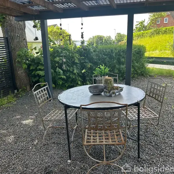 Et rundt bord med fire metalstole står under et halvtag. Bordet er dekoreret med planter, og omgivelserne er præget af grus og grøn beplantning.