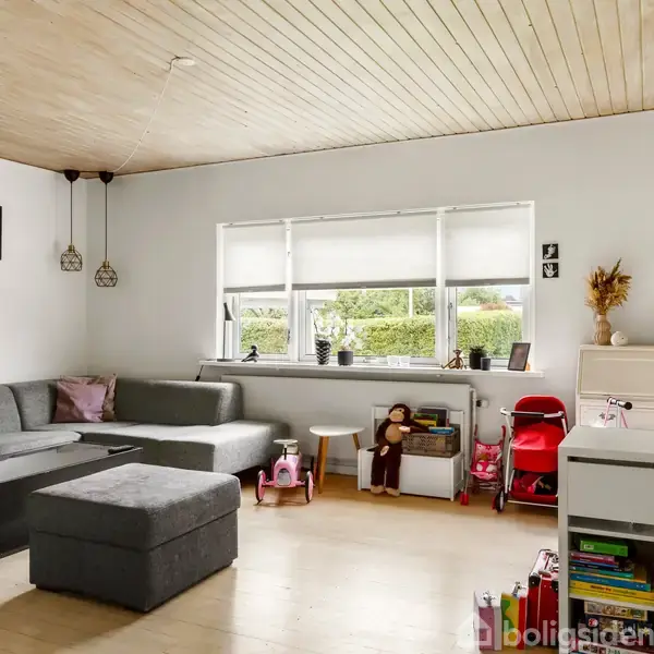 Gråt hjørnesofaarrangement står stille i en lys stue. Væggene prydes af små billeder, og legetøj er placeret nær vinduet. Et skrivebord står til højre for sofaområdet.