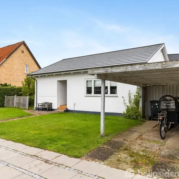 En hvid, enkelthus med gråt tag har en carport med en cykel. Det er omgivet af en grøn græsplæne og står ved siden af en stenlagt sti.