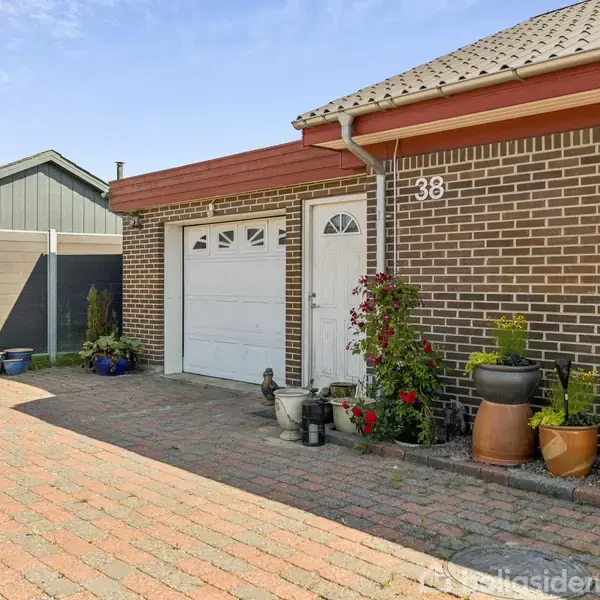 Husets facade består af mursten med en hvid garageport og grønne planter i krukker foran. Nummeret "38" er synligt. Omgivet af en flisebelagt indkørsel og hegn i baggrunden.