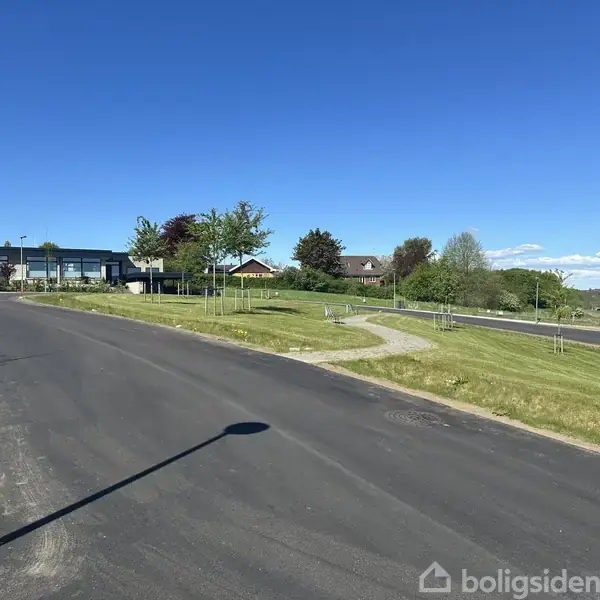 Nylagt asfaltvej strækker sig gennem et parcelhuskvarter med græsplæner og nyplantede træer. Enkelte moderne huse ses i baggrunden under en klar blå himmel.