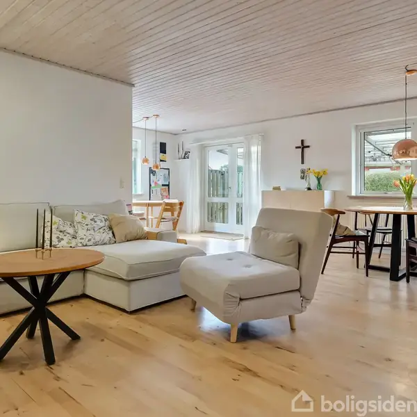 Sofaen står i en stue med lyst trægulv og hvidt loft. Den vender mod et rundt bord, mens spisebordet bagved har blomster og udsigt til en have gennem store vinduer.