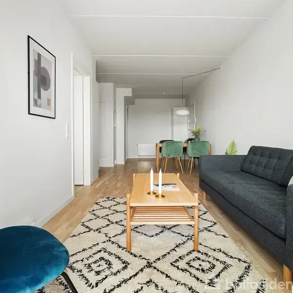 Sofa og stol i stue; sofaen er sort med puder, mens stolen er blå. Foran står et træbord med lysestager på et mønstret tæppe. I baggrunden ses et spisebord med stole.