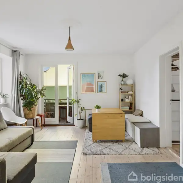 Stue med sofa og sofabord, kig til altan gennem glasdør, planter i potter ved vinduet, enkeltstående lampe i loft, lyse vægge og trægulv, indgang til tilstødende rum.