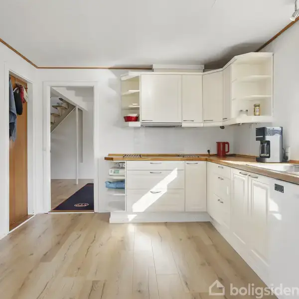 Et lyst, rummeligt køkken med hvide skabe og træbordplader. En kaffemaskine står på bordet ved siden af en vask. En dør fører til en trappeopgang i baggrunden.