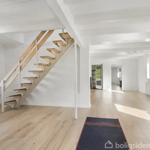 Trætrappe fører opad i et lyst rum med lyse trægulve og hvidmalede vægge. Store vinduer slipper naturligt lys ind, mens en dør leder til et tilstødende rum. En sort måtte ligger på gulvet.