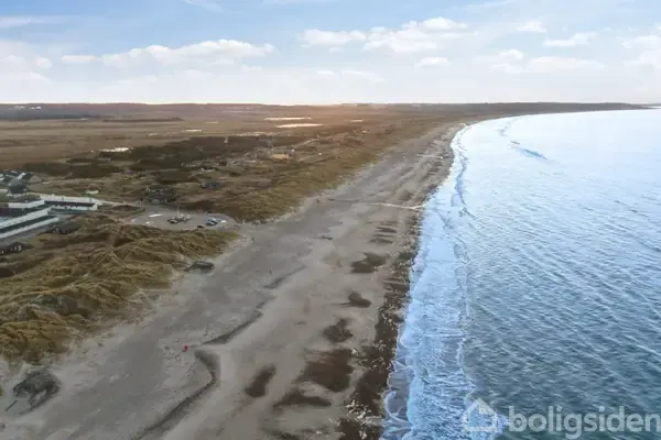 Bygninger ligger stille, malet hvidt og med skrånende tage, i et åbent, græsklædt kystlandskab under en klar, blå himmel.