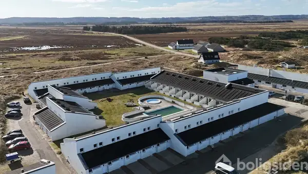 Køkken står med rummelige hvide skabe og skuffer, en kaffemaskine, og en komfur på en flisegulv. Området er lyst og rent, med træloft og hvidmalede vægge.