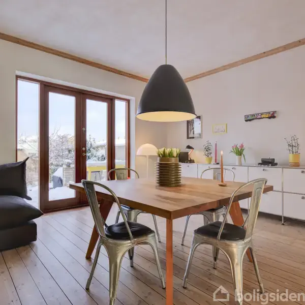 Et spisebord med metalstole står i en stue. En sort lampe hænger over bordet. Store vinduer åbner ud til en terrasse. Kommoden bagerst har dekorationer.