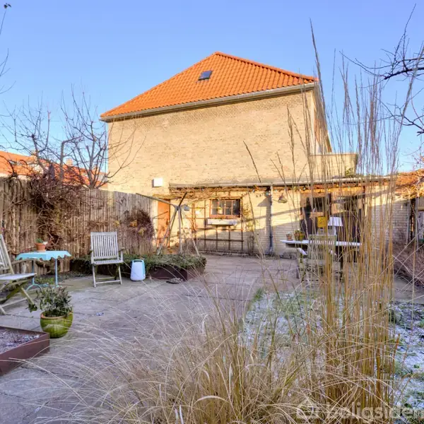 En gårdhave med to træstole og et bord står på en flisebelagt terrasse. Omgivet af højt græs og et hegn, ses et hus med rødt tag i baggrunden.