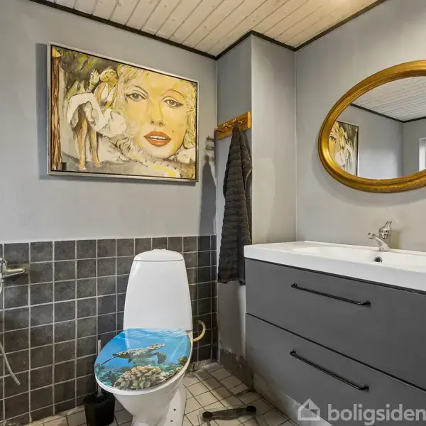 Et badeværelse med et toilet, hvis låg har et havtema. Over vasken hænger et ovalt spejl. På væggen er et farverigt maleri af et ansigt.