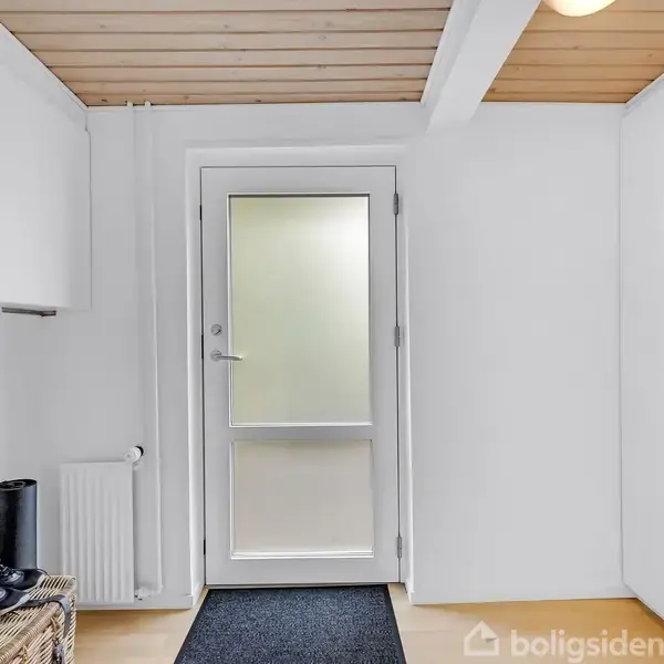 Dør med matteret glas står i en lys entré med træloft. En skohylde og en sort måtte ligger på gulvet. Vægge er hvide med en radiator og et skab.