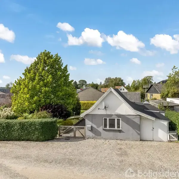 Et lille, hvidt hus med gråt tag står stille i en rolig, hvidmalet boligområde. Omgivet af velplejede hække, træer og grusvej under en blå himmel med hvide skyer.