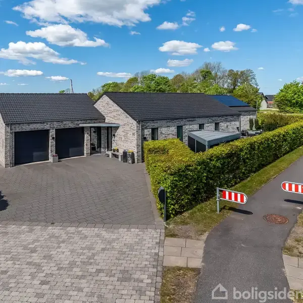 Et moderne hus med en grå murstensfacade og sort tag står stille ved siden af en asfalteret sti, omgivet af velholdt hækbeplantning under en klar, blå himmel.