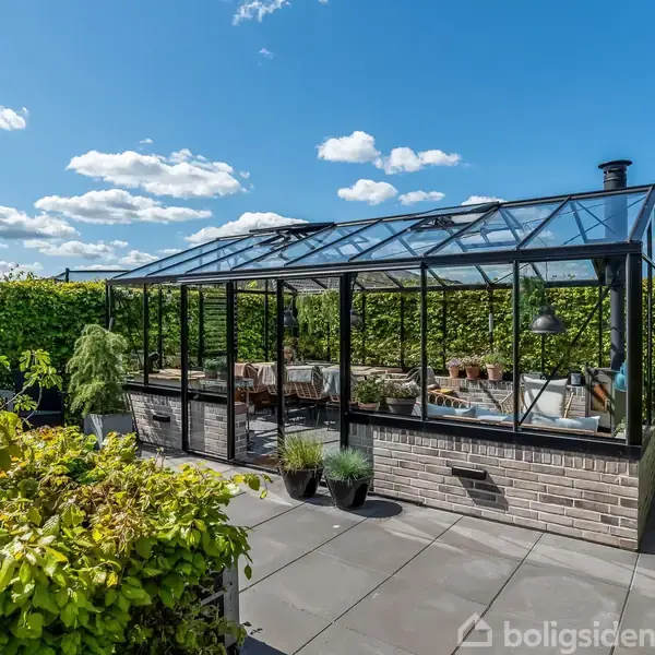 Et drivhus med glasvægge står på en flisebelagt terrasse, omgivet af grønne planter og hække, under en klar blå himmel med let spredte skyer.