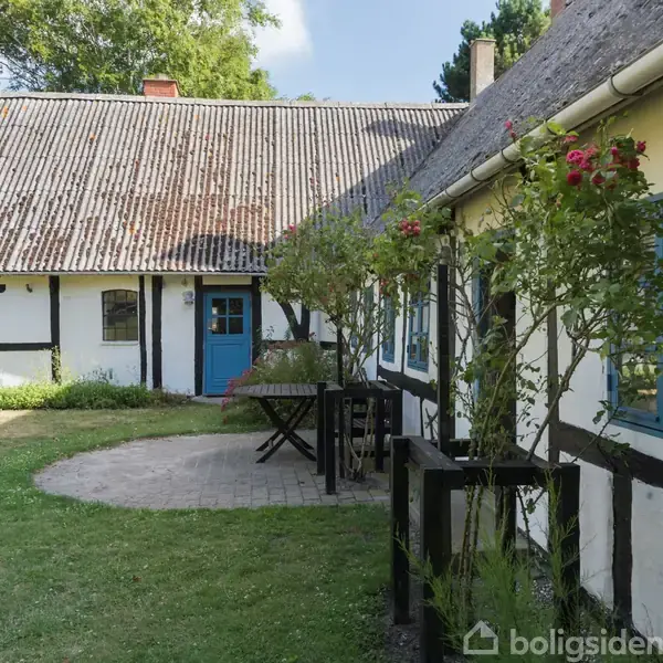 Bindingsværkshus med sort-hvid facade står i en grøn have. Bluemalet dør og vinduer tilføjer kontrast. Et bord med stole står på en flisebelagt cirkel foran huset.