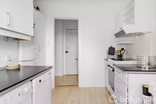 Et lyst køkken med hvide skabe og apparater; opvaskemaskine til venstre, komfur og emhætte til højre; et snorlige trægulv og en åben dør til en entré.