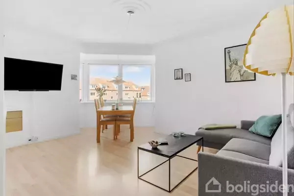 Sofa står i en stue med et sofabord foran. Et spisebord med stole er ved et vindue i baggrunden. En fladskærm hænger på væggen til venstre.