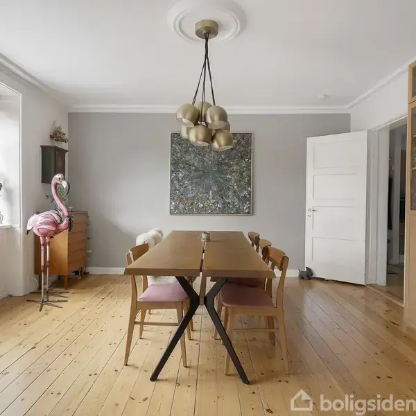 Et rektangulært træbord står i midten af et lyst rum med lyse trægulve. Seks stole omgiver bordet, en kunstnerisk lampe hænger over det, og en flamingoskulptur står ved vinduet.